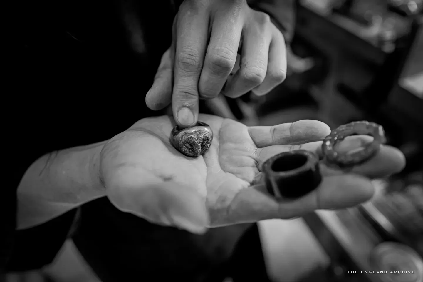 A hand pinches a textured cast ring on an open palm, two more rings - a plain band and a filigree band - resting in the palm.
