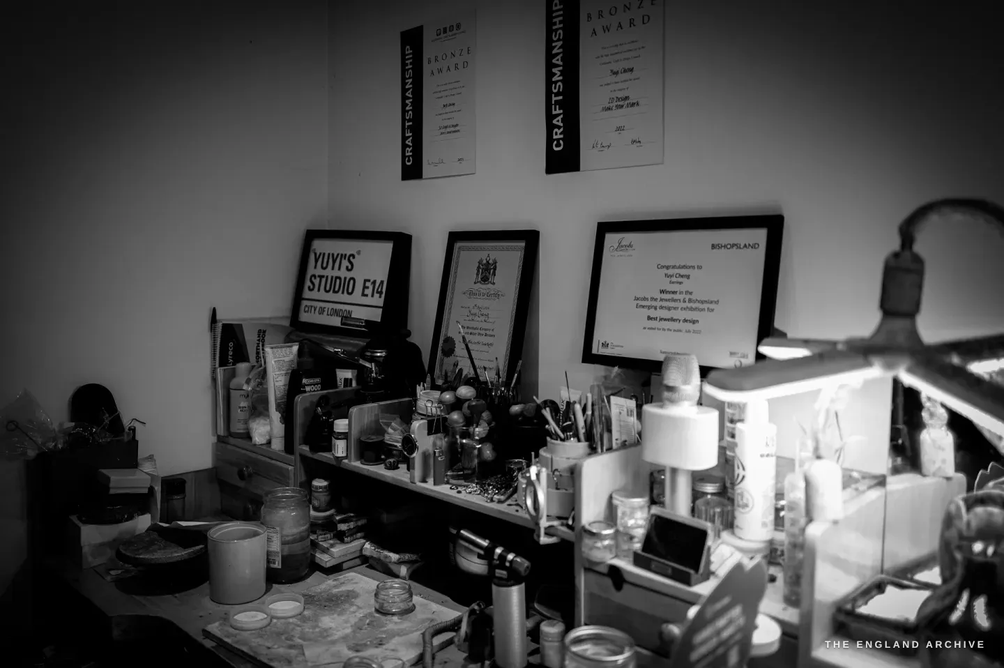 The wall above the bench: framed certificates and two "Bronze Award - Craftsmanship" banners, a "Yuyi’s Studio E14" sign, the bench below crowded with brushes, jars and tools.
