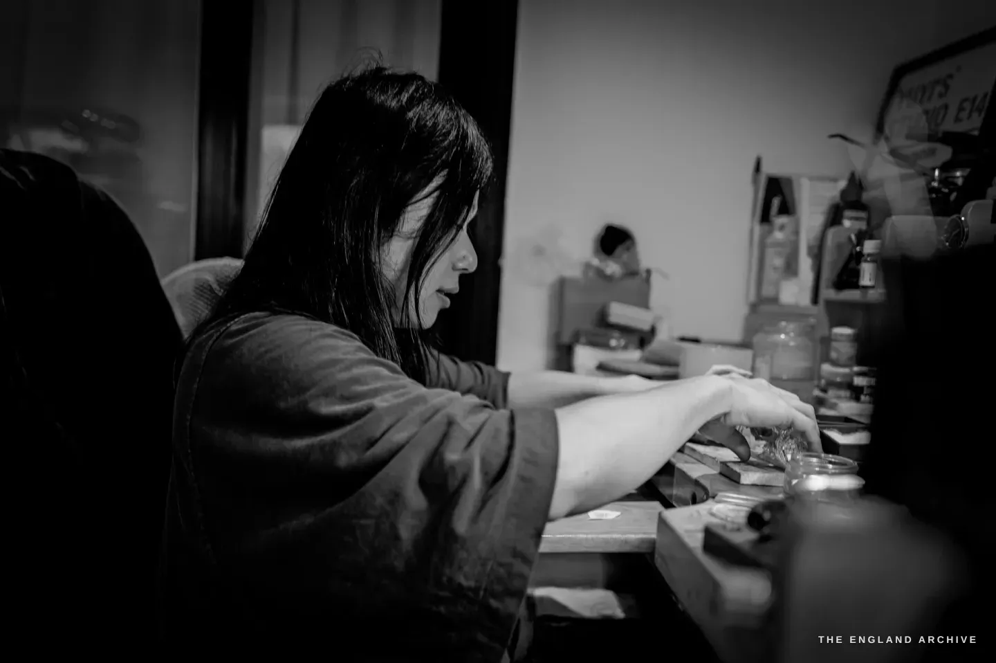 Yuyi in profile, leaning over the bench, both arms reaching forward into jars and tools at work.