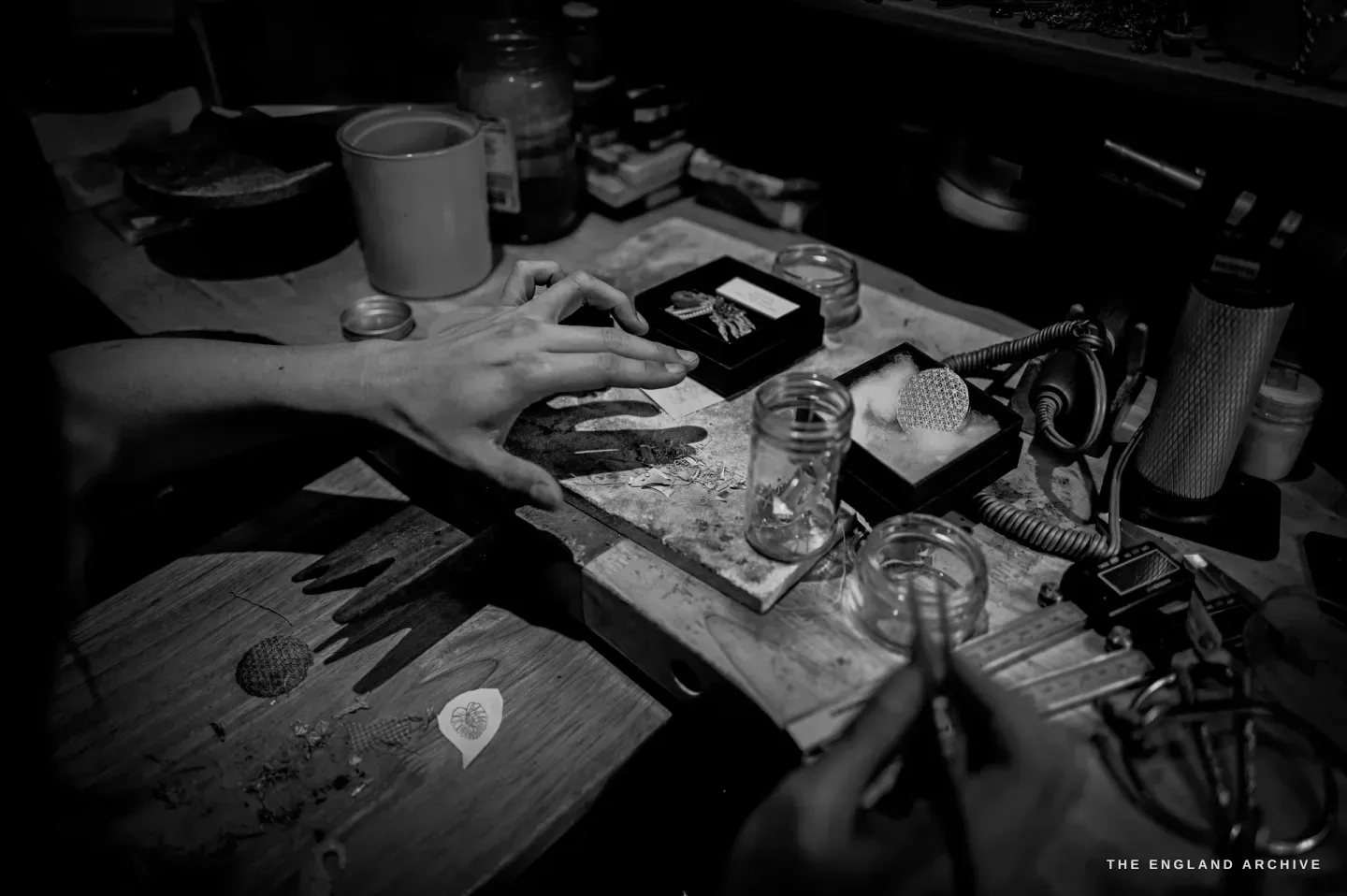 Over-the-shoulder of the bench: a hand by a black box holding a brooch and a label, jars of fluid, a circular openwork filigree disc in a tray, a soldering airline.