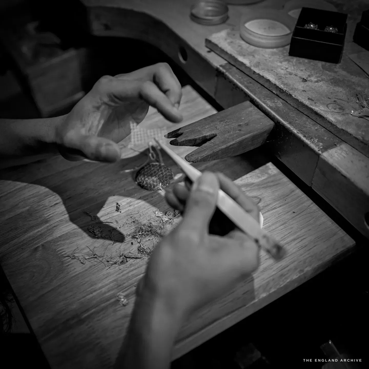 Close over-the-shoulder view of two hands working a fine brush over the bench pin, a domed openwork filigree sphere and silver wire offcuts on the peg.