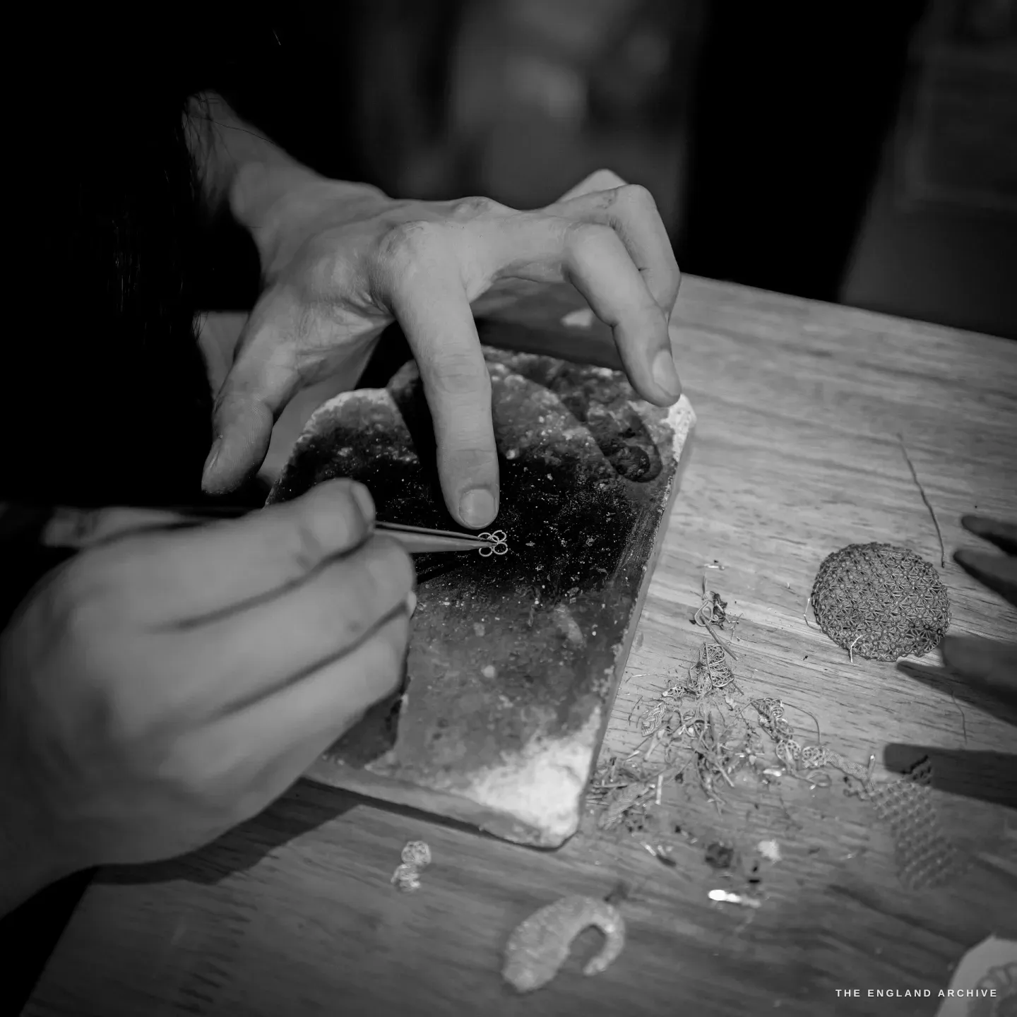 Hands using fine tweezers to set tiny filigree scrollwork against a charcoal soldering block, a hemispherical openwork filigree dome and offcuts of silver wire on the bench.