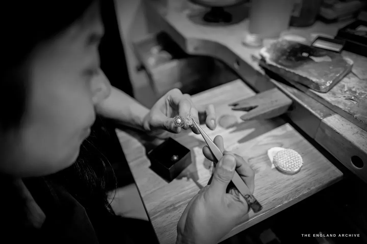 Over-the-shoulder: Yuyi holds tweezers gripping a pair of small filigree flower-and-pearl earrings, a round filigree disc and a ring box on the bench.