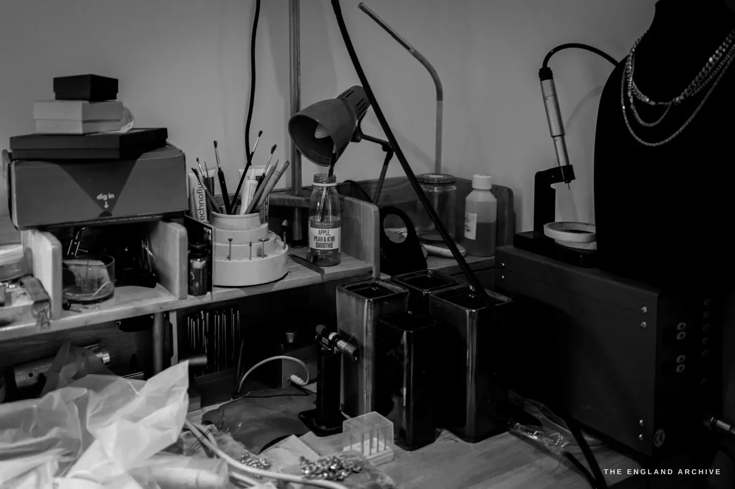 A bench-side corner with a desk lamp, a jar of blue and white chips, a butane torch, dark ingot moulds and a polishing pendant motor against a black necklace bust.