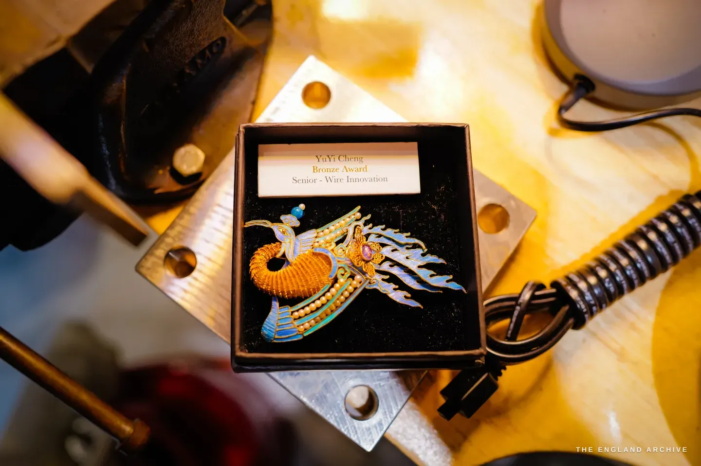 A presentation box on the bench holding a gold-and-blue feather phoenix brooch set with seed pearls, a steel vice and soldering airline framing it; the box label reads "YuYi Cheng, Bronze Award, Senior - Wire Innovation".