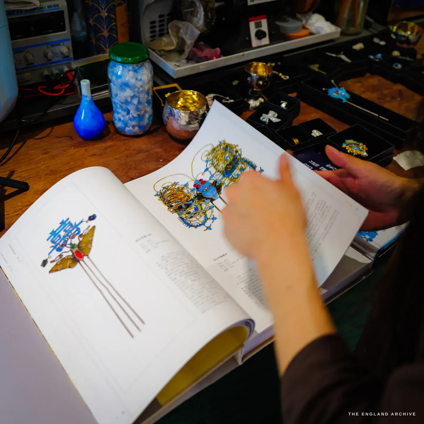 A hand turns the pages of a Chinese-language reference book showing antique blue dian cui filigree headdresses, surrounded by jars, a silver cup and trays of her own pieces.