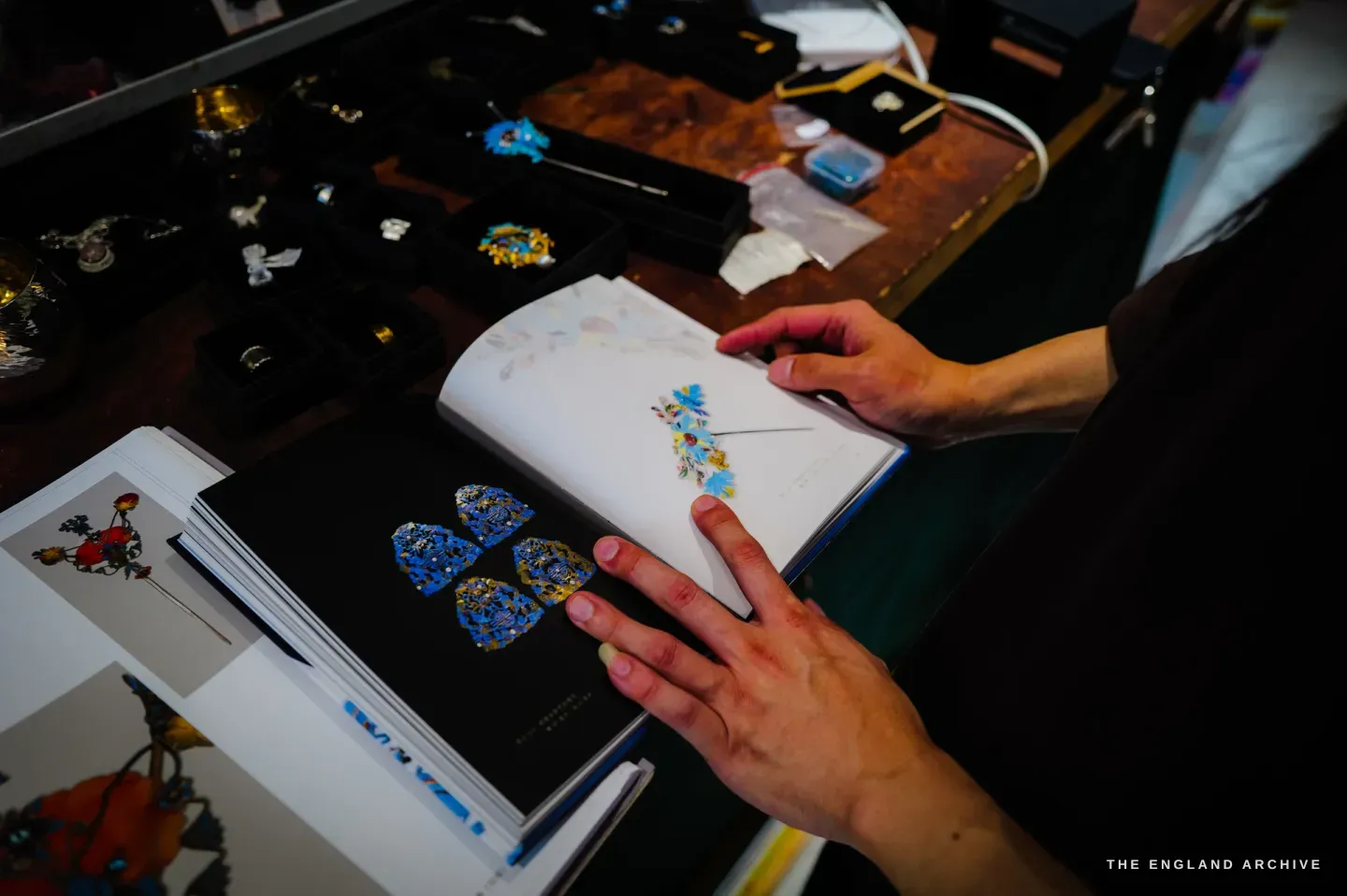 Yuyi holds open a book showing blue-and-gold filigree ornaments on a black ground, her finished pieces in trays behind.