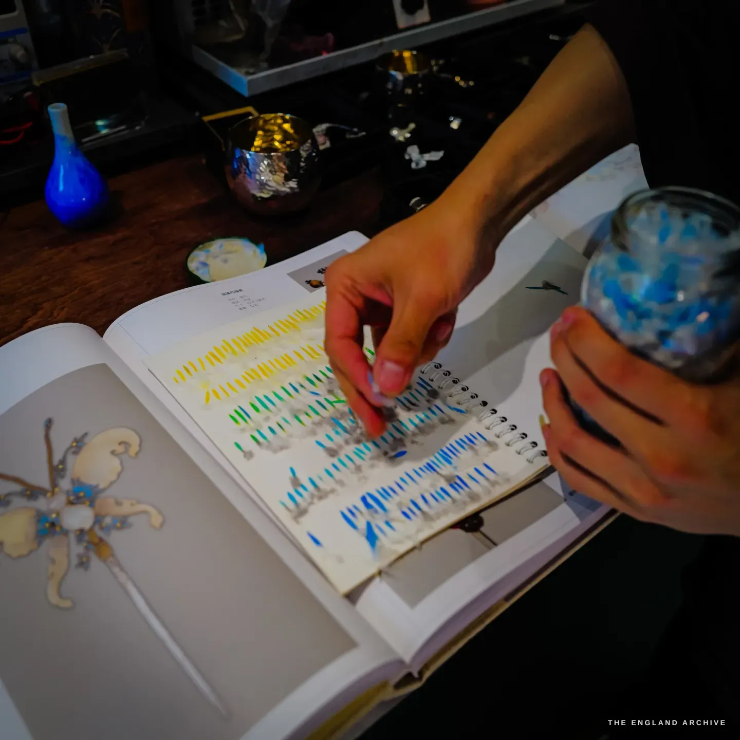A hand turns a spiral-bound album page filled with graded rows of cut feather sections, from yellow through green to blue, holding a small jar of blue feather chips.