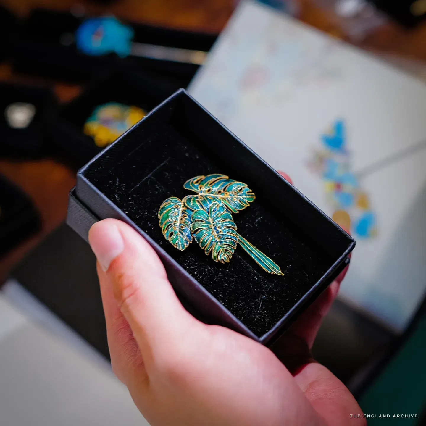A hand holds a black box containing a three-leaf green-and-gold dian cui monstera-leaf brooch.