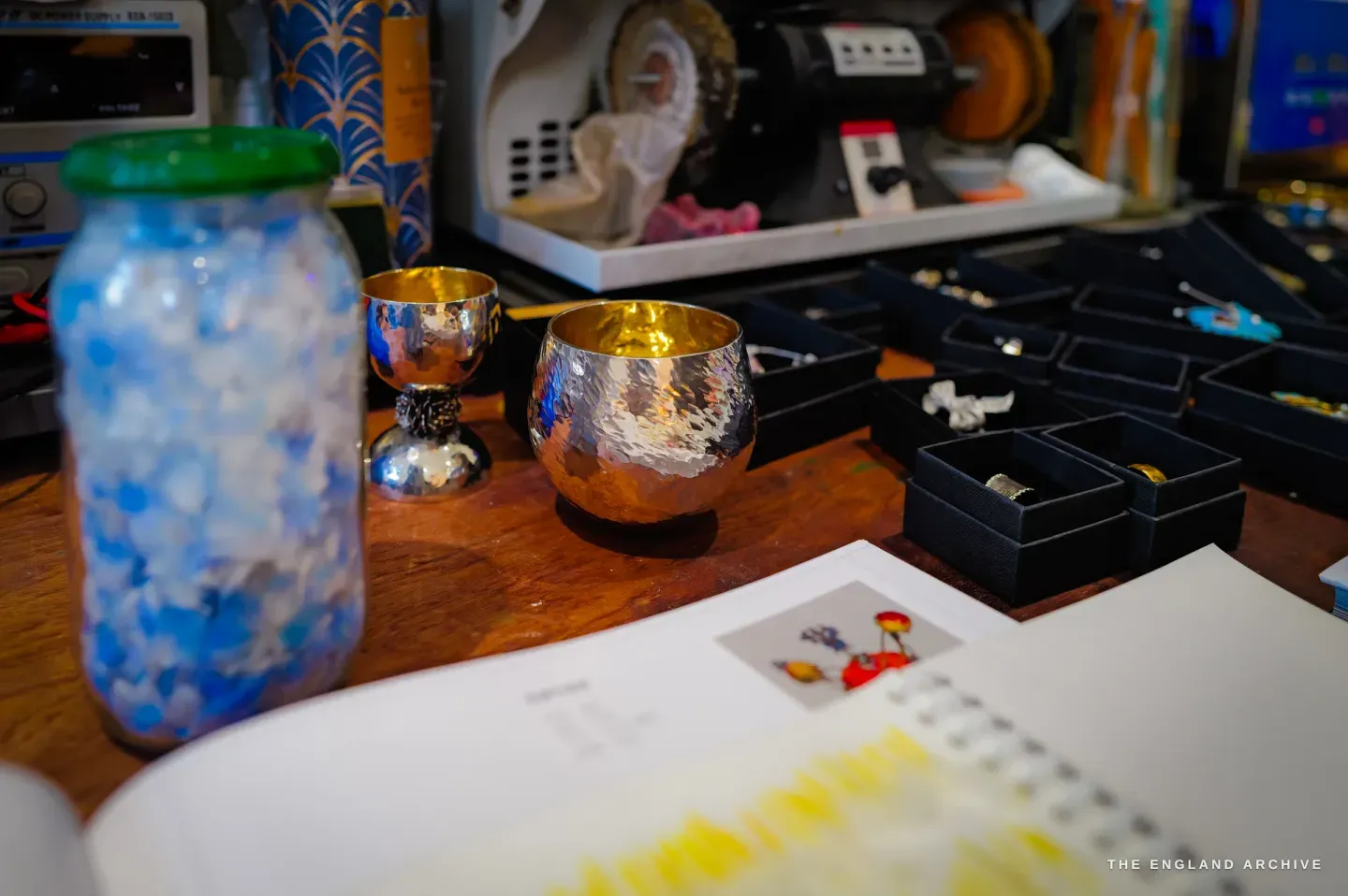 A bench still life: a jar of blue and white chips, two hammered silver-and-gold cups in a sake-cup form, a polishing motor behind, trays of rings to the right.