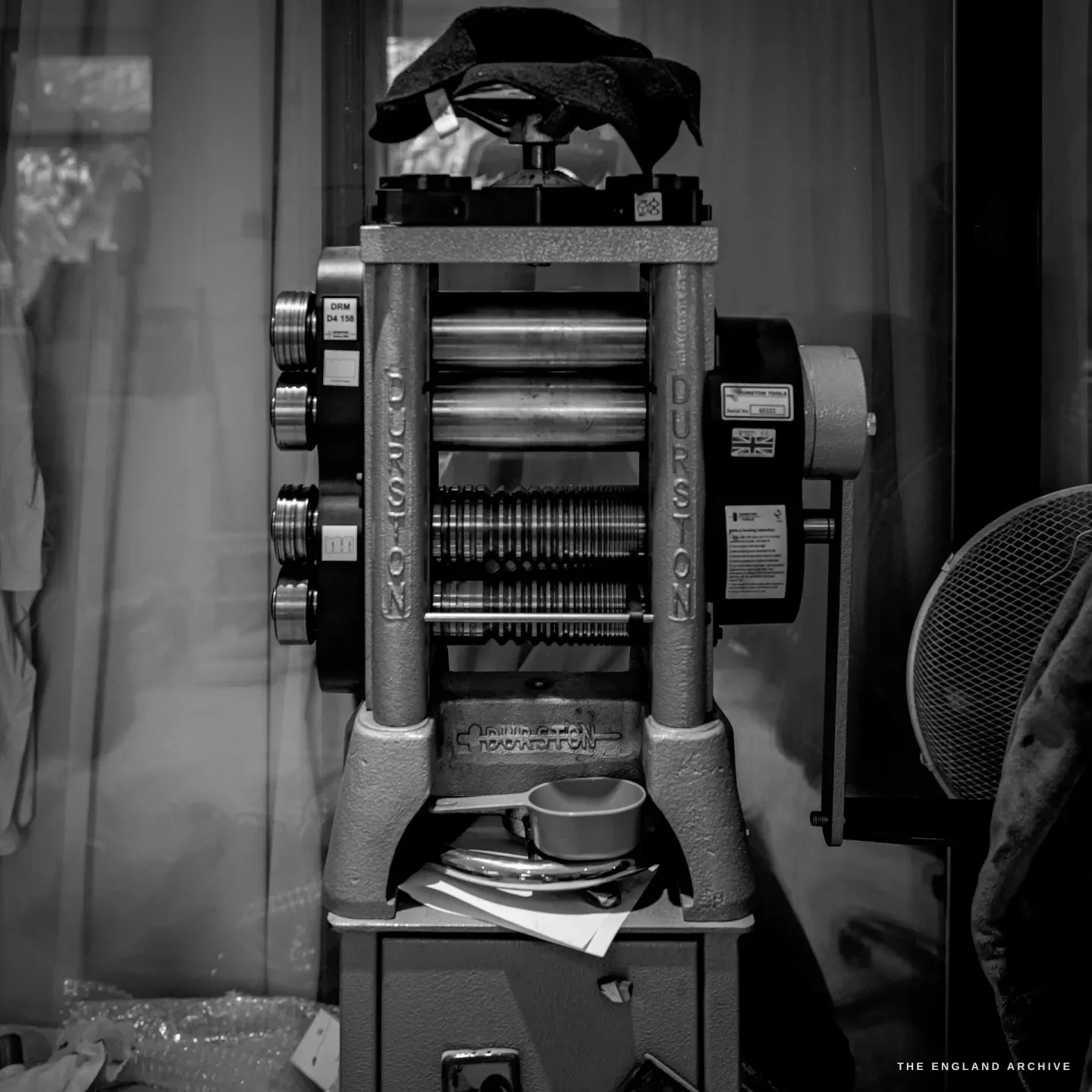 A hand-cranked Durston rolling mill standing by the window, a cloth draped over it, an electric fan beside it.