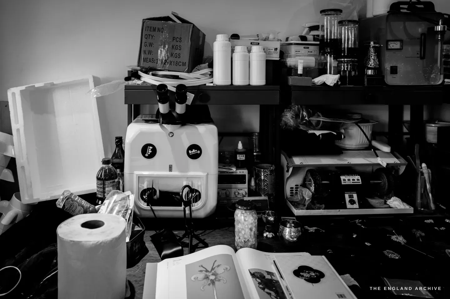 An equipment corner: a laser spot welder with its binocular viewing head on a stand, white reagent bottles, a bench lathe on a pull-out shelf, an open reference book of dian cui ornaments in the foreground.