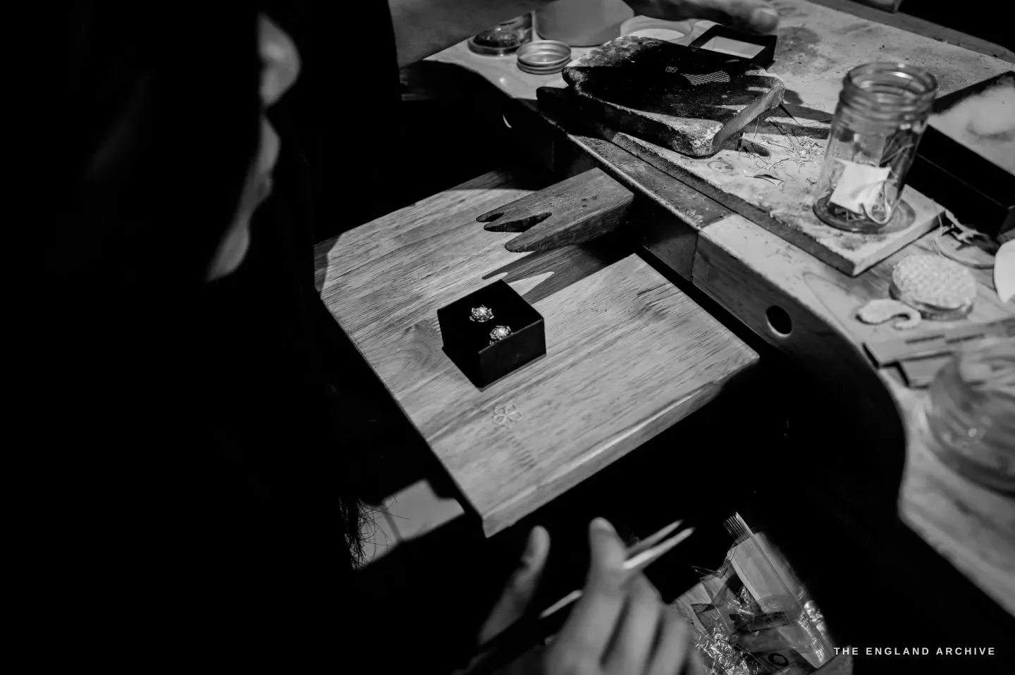 Looking down to a wooden board where a black box holds a pair of filigree pearl flower studs, a hand with tweezers reaching in, a soldering block and a jar of wire to one side.