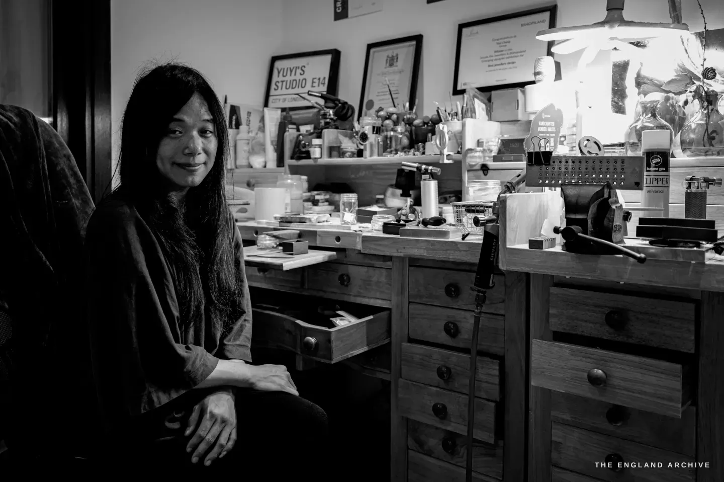 Yuyi Cheng seated beside her full jeweller’s bench, hands folded, smiling softly at the camera, framed awards and a hand-lettered "Yuyi’s Studio E14" sign on the wall behind her.