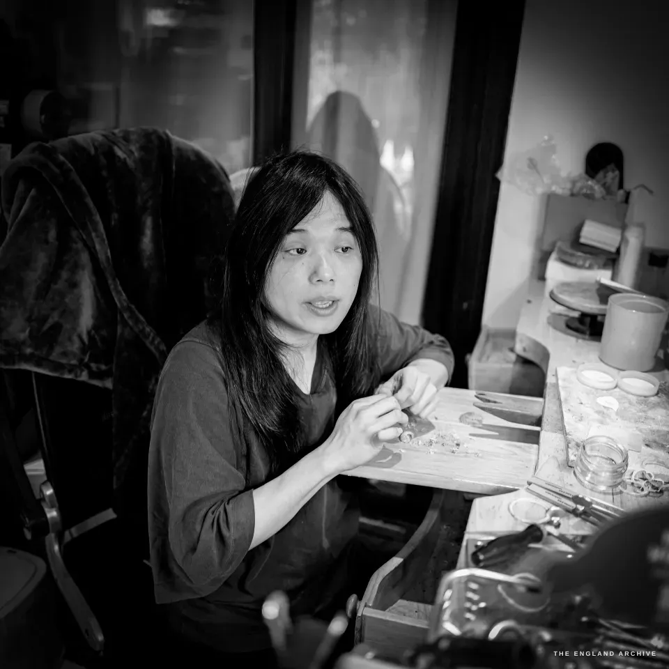 Yuyi at the bench mid-speech, looking off-camera, one hand working over the bench pin, jars and tools around her.