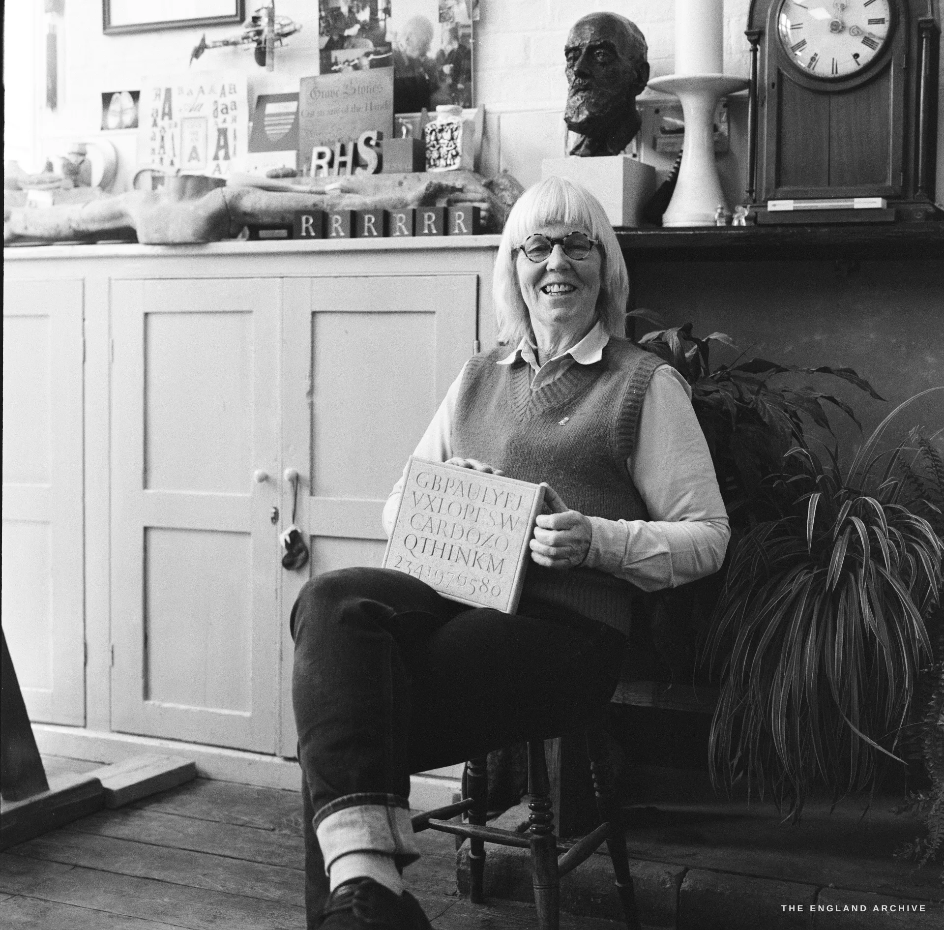 Lida Kindersley seated in the lettering workshop, holding a carved slate that reads CARDOZO, smiling at the camera, plaster busts and a clock on the shelf behind.