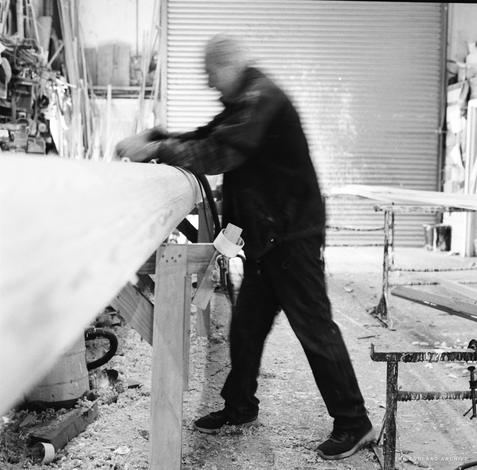 A motion-blurred film frame of Michael Dennett working a long timber spar at the boat-builder's bench, his upper body and arms smeared by a slow shutter.