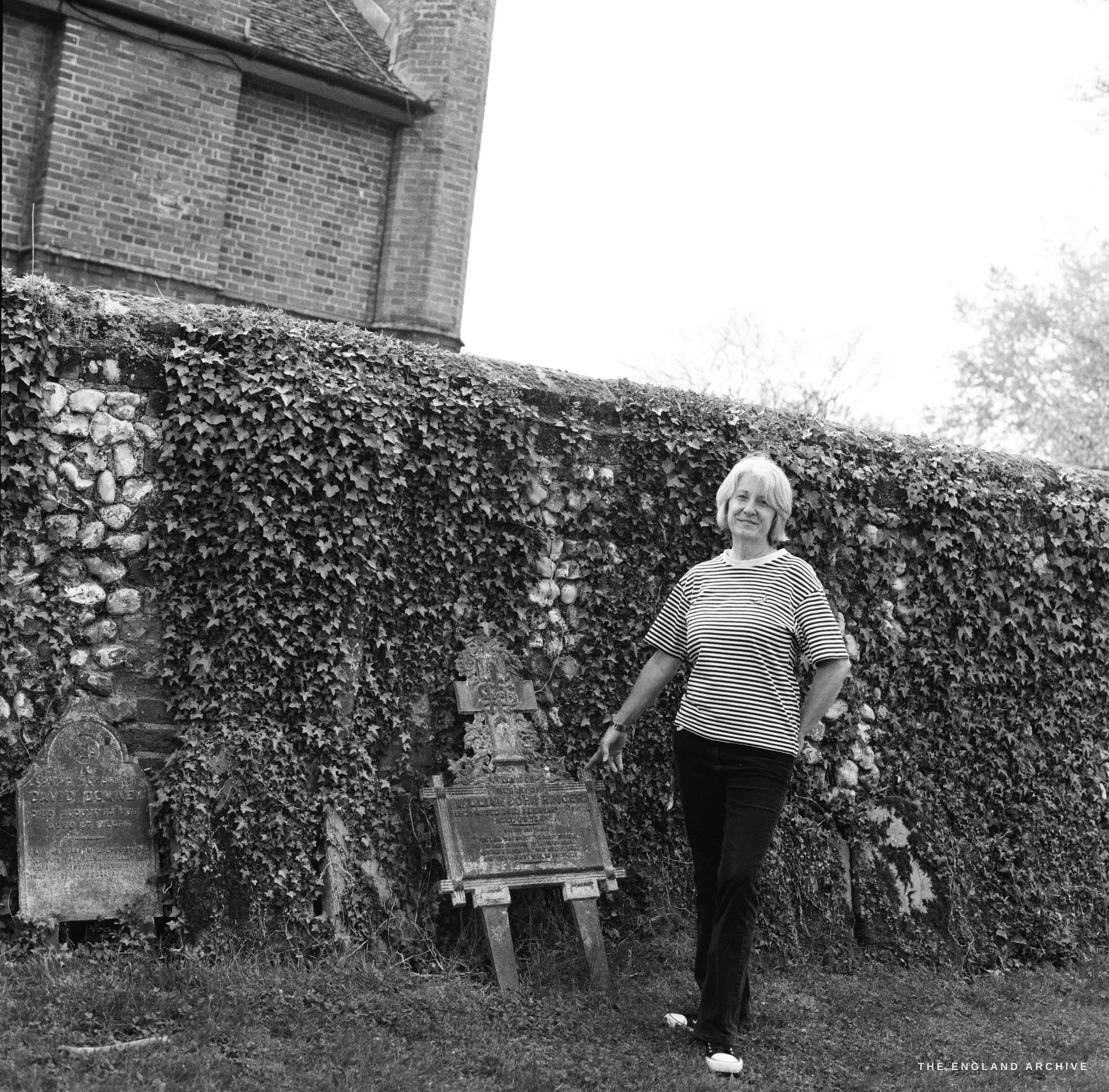 A film portrait of a woman in a striped top standing beside an ivy-covered flint churchyard wall at Long Melford, gravestones to her left.