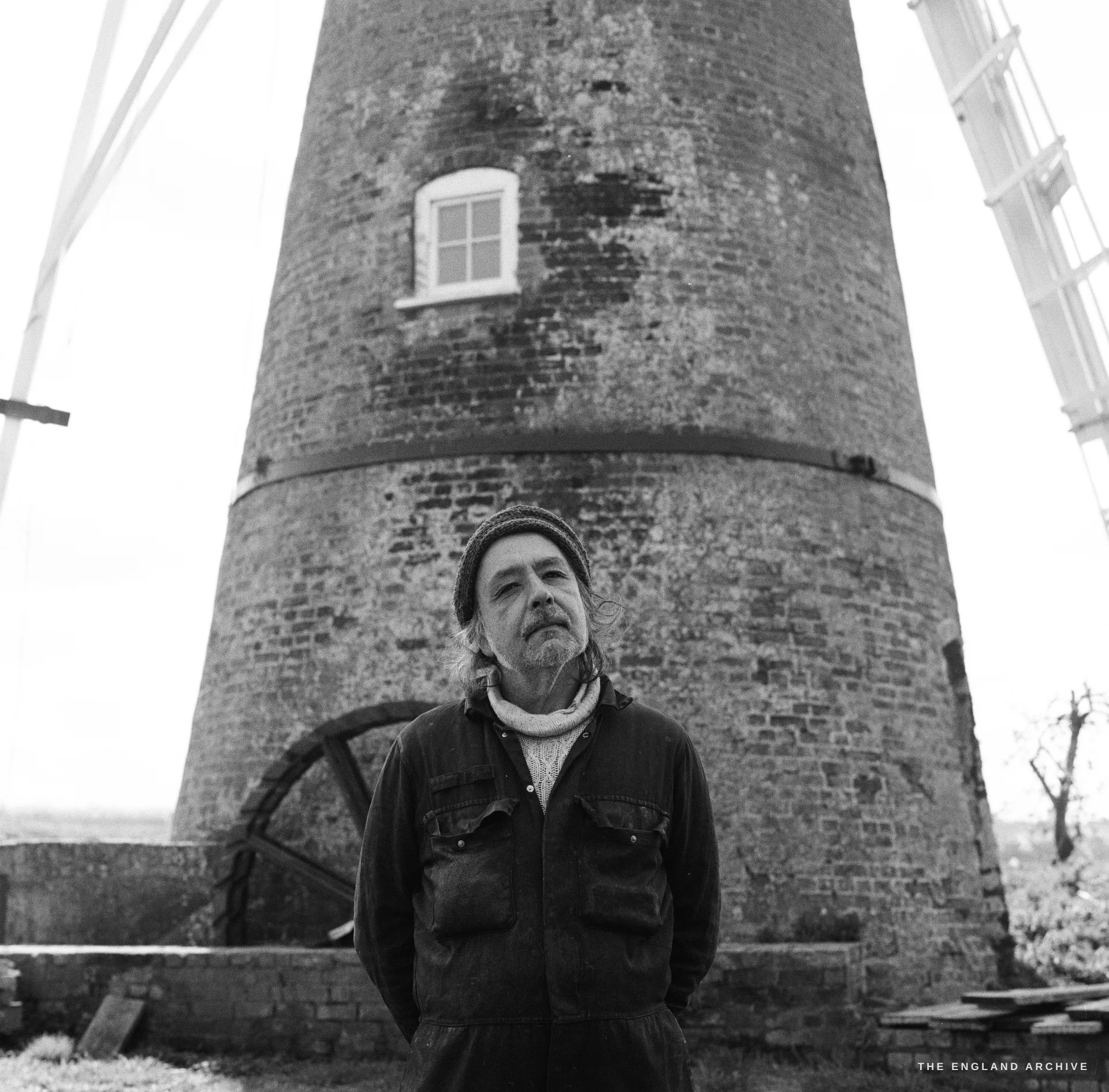 Paul Kemp standing at the brick base of a tower windmill, sails overhead, looking up at the camera, the flat marsh behind.